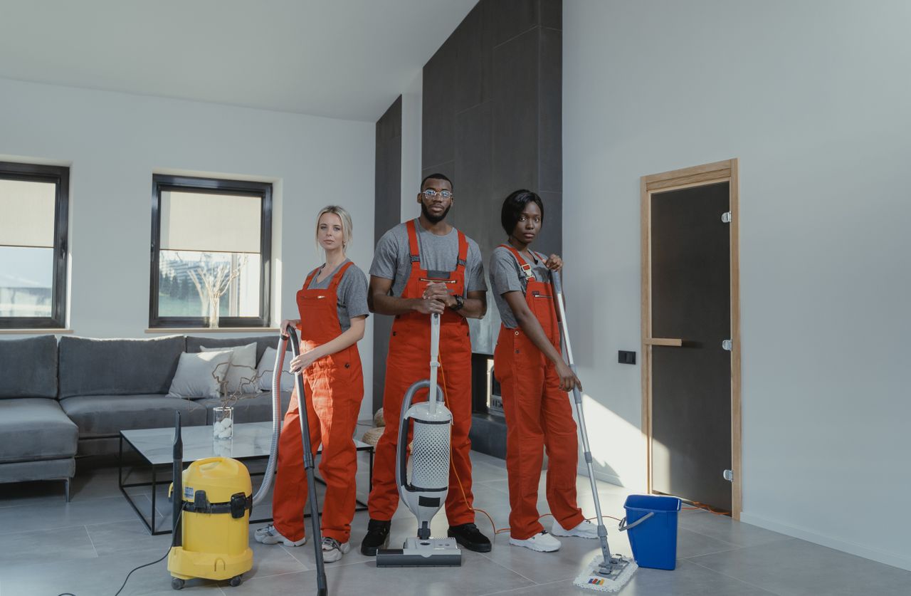 Hello world! Diverse cleaning team in red uniforms ready to sanitize a modern living room with vacuum and mop.