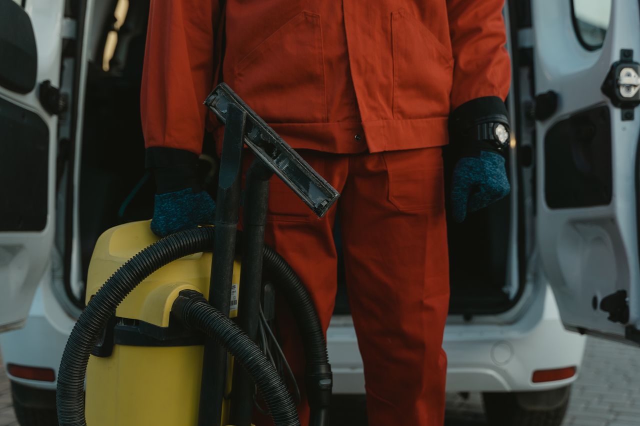 who-we-are A professional cleaner in a red uniform holding a vacuum near a van, ready for service.